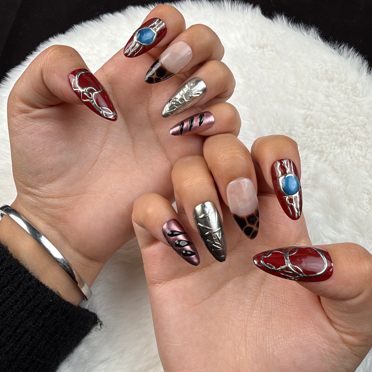 Hands wearing Velvet Relic burgundy and silver chrome almond press-on nails with a blue stone accent