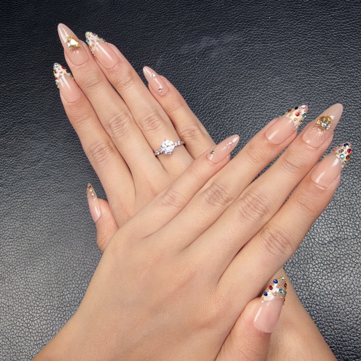 Hands wearing Golden Heart peach nude jewel almond press-on nails with a gold heart and crystals