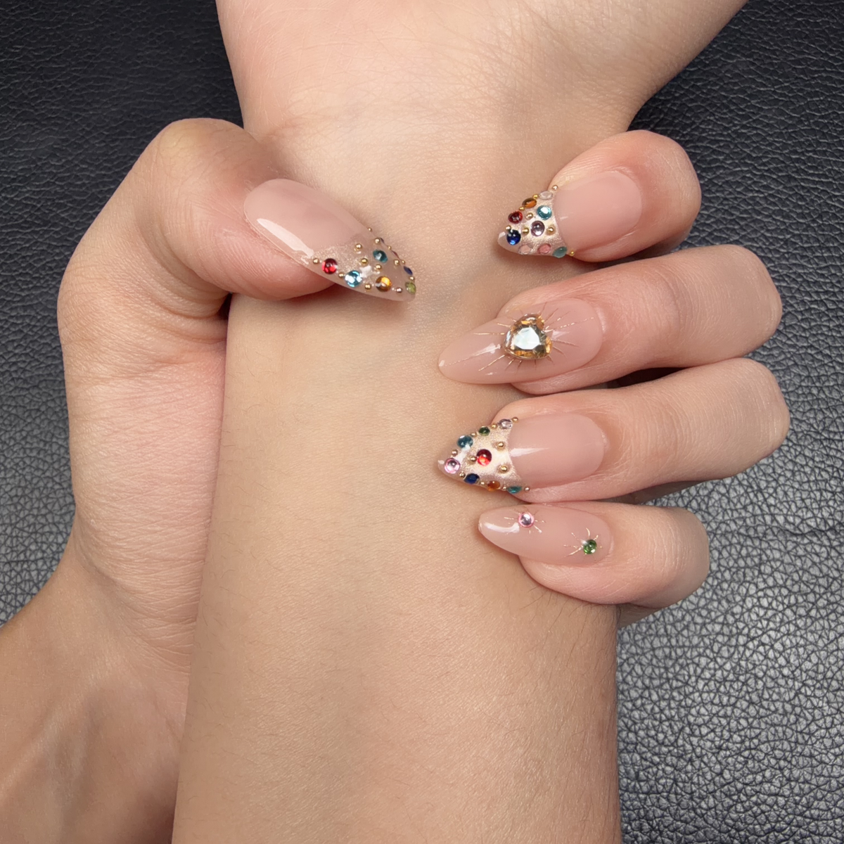 Hands wearing Golden Heart peach nude jewel almond press-on nails with a gold heart and crystals