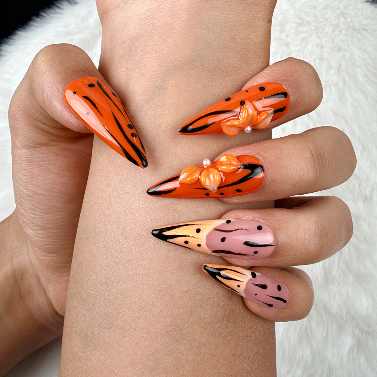 Detail shot of Tangerine Edge orange chrome and nude pink stiletto press-on nails with black floral details on hand