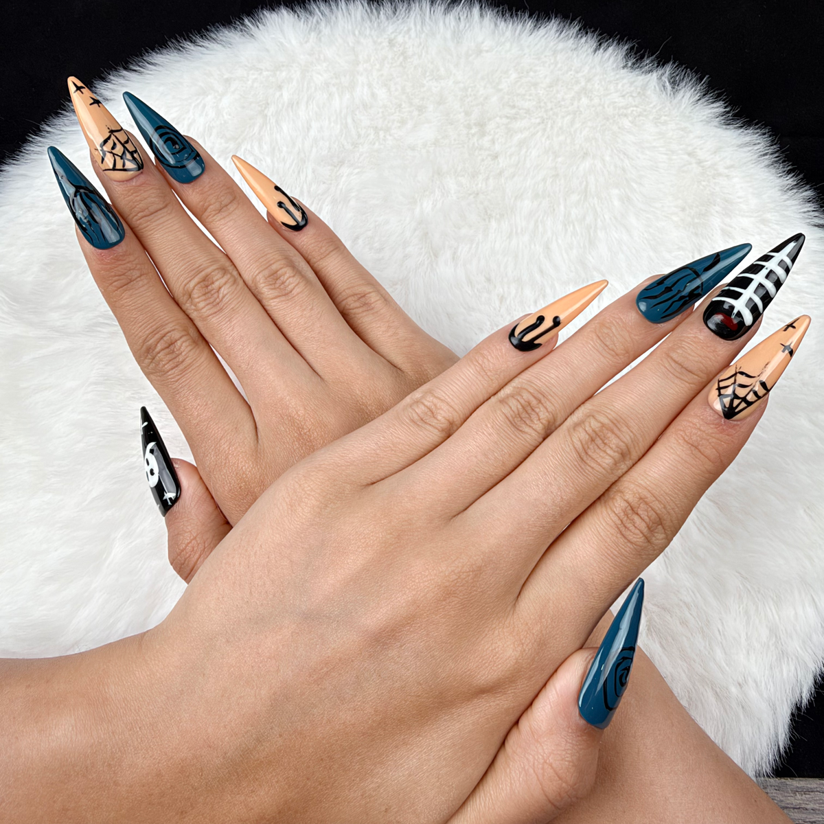 Hands wearing Twilight Web teal, black and peach Halloween stiletto press-on nails with skulls and spiderwebs