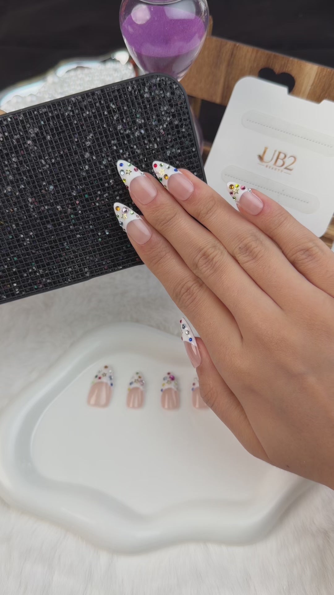 Load video: Step-by-step application of White French tip almond press-on nails with colorful rhinestones and star accents